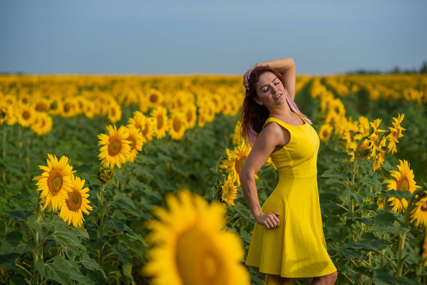 A red-haired woman in a yellow dress is standing in a field of sunflowers. Beautiful girl in a skirt sun enjoys a cloudless day in the countryside. Pink locks of hair. - Фото, зображення