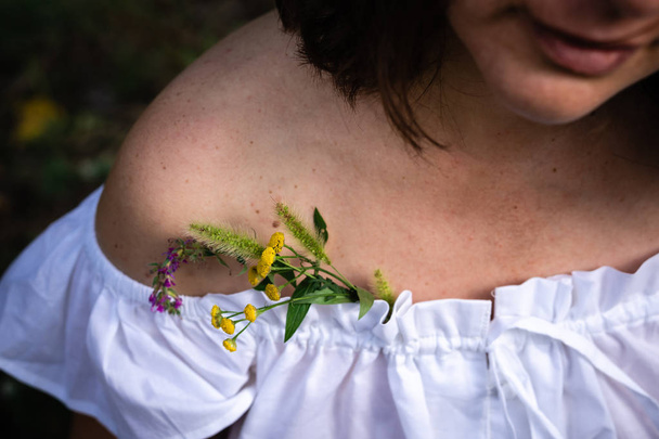 ein kleiner Strauß Wiesenblumen versteckt in einem weißen Hemd auf den nackten Schultern einer Frau. heller Ton. horizontale Ausrichtung - Foto, Bild
