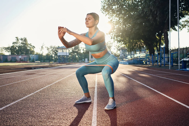 Beautiful sporty girl in stylish sportswear squatting with rubber band during workout on city stadium - Zdjęcie, obraz