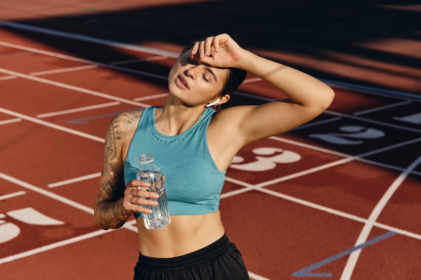 Спортсменка в спортивной одежде с бутылкой воды устало держась за руку возле головы после тренировки на городском стадионе
 - Фото, изображение