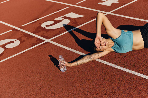 Aantrekkelijke atleet meisje in sportkleding liggend op runner track dreamily bedekken ogen met de hand van zon na training op stadion - Foto, afbeelding