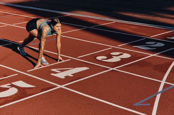 Привлекательная спортсменка в спортивной одежде, стоящая в стартовой позиции, готовая к бегу по городскому стадиону
 - Фото, изображение