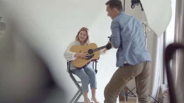 Színfalak mögött a fotózásra. Híres szakmai fotós fotózni a fiatal nő gitározni, miközben ül a széken, fehér háttér a stúdióban. Divatstúdió fotózás. - Felvétel, videó