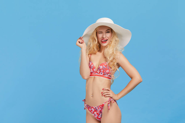 Portrait of a smiling and shocked attractive woman in bikini and hat points a hand to the right and up and looking at camera isolated over blue background. Travel and vacation concept.  - Foto, Bild