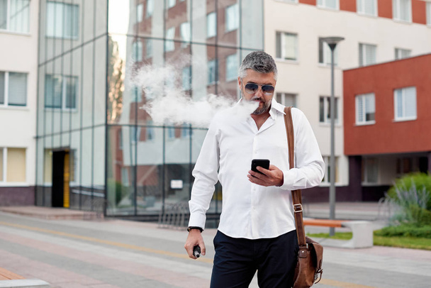Middle-aged bearded thinking serious concentrated business man standing and smoking electronic cigarette. Blows smoke through the mouth. In-ear wireless earphone. Office centre background - Fotografie, Obrázek