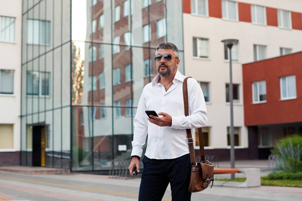 Middle-aged bearded thinking serious concentrated business man standing and smoking electronic cigarette. Blows smoke through the mouth. In-ear wireless earphone. Office centre background - Foto, Bild