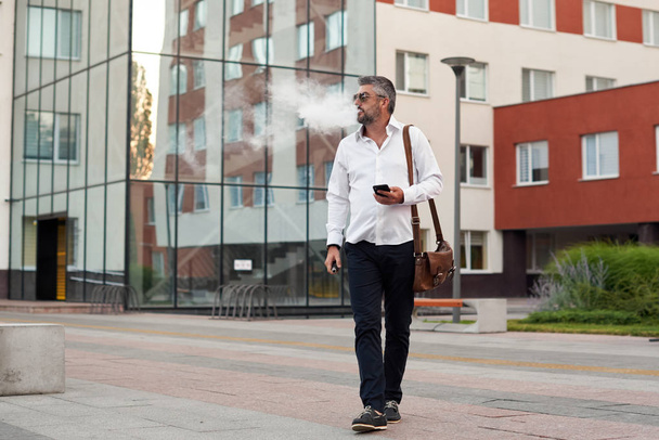 Middle-aged bearded thinking serious concentrated business man standing and smoking electronic cigarette. Blows smoke through the mouth. In-ear wireless earphone. Office centre background - Фото, изображение