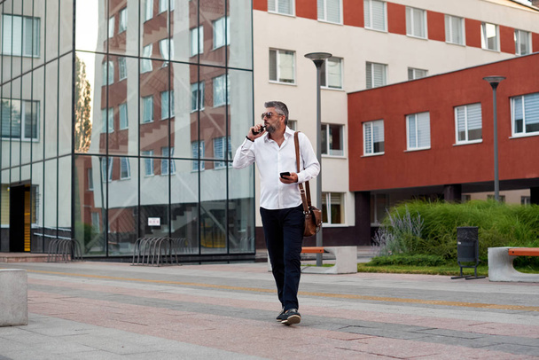 Middle-aged bearded thinking serious concentrated business man standing and smoking electronic cigarette. Blows smoke through the mouth. In-ear wireless earphone. Office centre background - Foto, imagen
