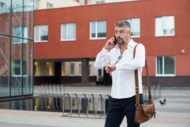 Middle-aged male businessman talking loudly on mobile phone and smoking a vape, blowing smoke. City and office building background - Foto, Imagem