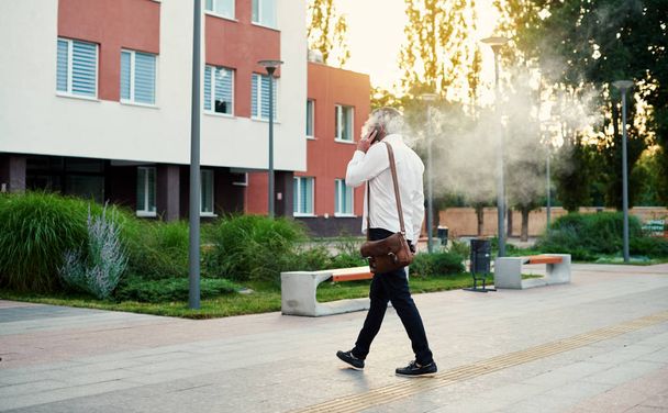Middle-aged male businessman talking loudly on mobile phone and smoking a vape, blowing smoke. City and office building background - Photo, Image