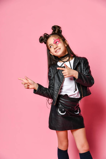 Little - fashionable hipster punk girl - dressed in a leather jacket and skirt, black knee-highs and a white T-shirt, with a funny hairstyle and a makeup star on the face, points to the side - Foto, Imagem