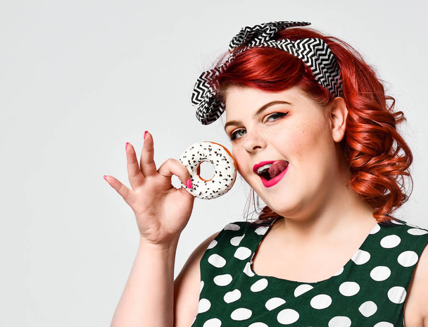 Portret van mooie vrolijke dikke plus size vrouw pin-up dragen van een polka-dot jurk geïsoleerd over lichte achtergrond, het eten van een donut - Foto, afbeelding