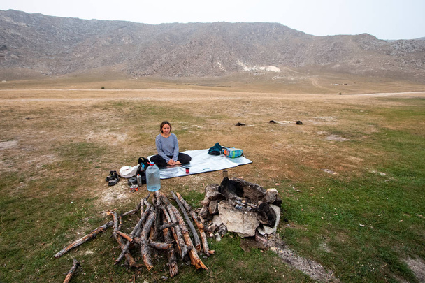 Girl tourist sitting in a yoga pose on a tourist silver rug by the fire. Behind the mountains. Nearby is a gas burner and a gas cylinder. Firewood has been prepared for the fire. The bonfire is lined with stones. Copy space. - Foto, Imagem