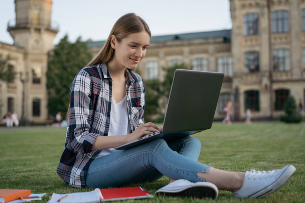 Sikeres egyetemi tanuló, tanuló nyelv a fűben ülve a parkban. Online oktatási koncepció. Gyönyörű mosolygó nő használ laptop számítógép, keresési információk, gépelés billentyűzet, munka projekt online - Fotó, kép