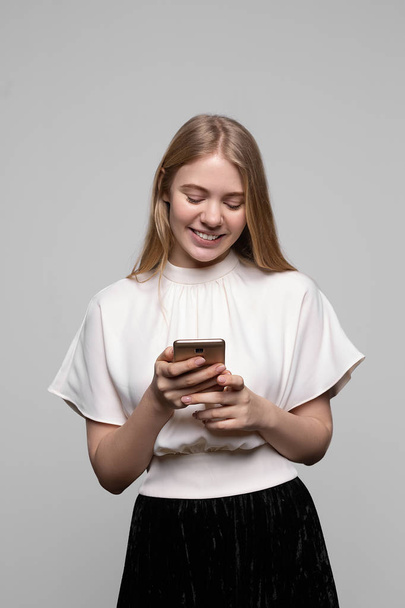 長い髪の笑顔と灰色の背景に隔離された携帯電話を使用して若いかわいいブロンドの女の子。現代のコミュニケーション - 写真・画像
