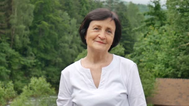Zbliżenie portret starszej kobiety z krótkimi ciemnymi włosami i pomarszczonej twarzy patrząc na aparat z uśmiechem na górskim wzgórzu z zielonym lasem na tle - Materiał filmowy, wideo