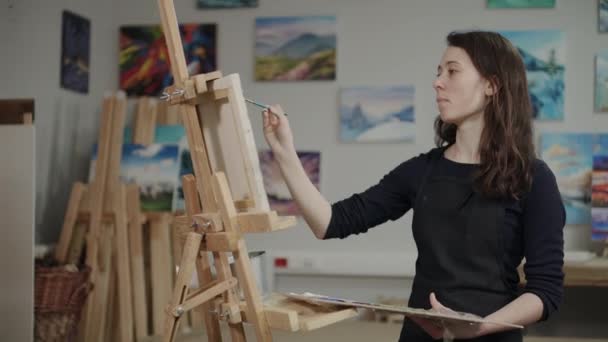 Νεαρή μελαχρινή γυναίκα δημιουργεί εικόνα σε καλλιτεχνικό εργαστήρι, στέκεται - Πλάνα, βίντεο