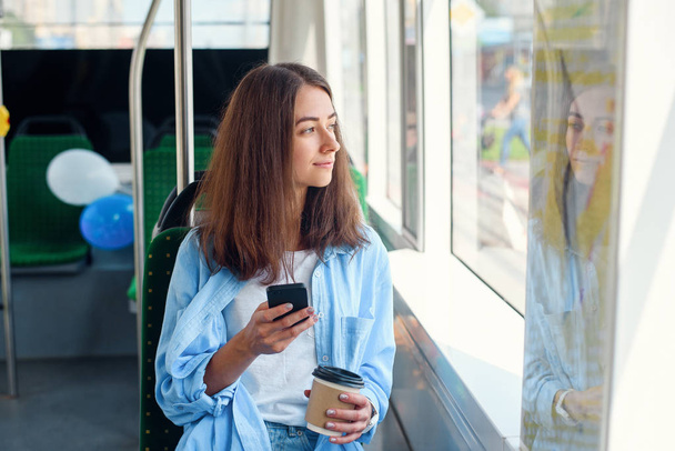 Pretty female passenger sits with smartp hone while moving in the modern tram or subway. Trip at the public transport. - Φωτογραφία, εικόνα