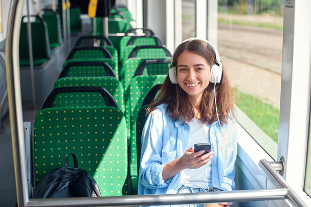 Pretty woman sits with headphones, smartphone and cup of coffee while moving in modern city tram. enjoying the trip at the public transport. - Foto, imagen