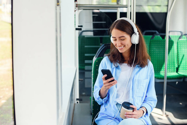 Young woman using public transport sits with smart phone and white headphones in the modern bus. - Fotografie, Obrázek