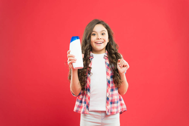 Hygiene habits for kids. Happy little girl holding gel or shampoo bottle for personal hygiene on red background. Small child with long hair smiling with hygiene product. Everyday hygiene and health - Photo, Image