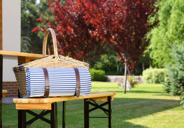 Picnic basket with blanket on wooden bench near table in green garden - Fotó, kép