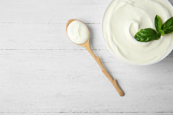 Bowl of fresh sour cream with basil and spoon on white wooden table, flat lay. Space for text - Foto, Imagen