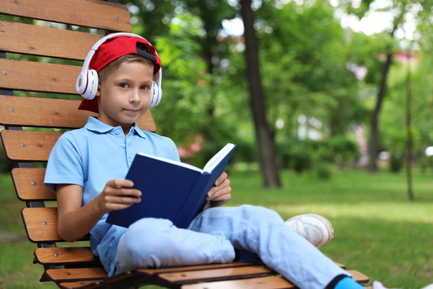 Cute little boy with headphones reading book on lounger in park - 写真・画像