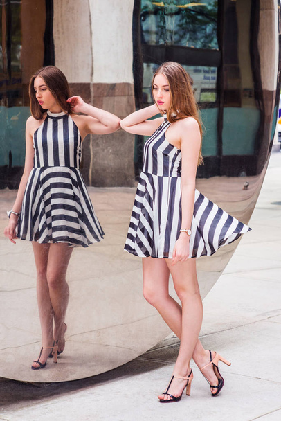 Young Beautiful Woman standing by metal mirror on street, looking down, thinking, wearing sleeveless, black and white striped short dress, sandals. Concept of self assured, self esteem, self checking - Photo, Image