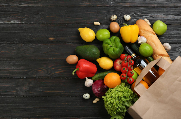 Flat lay composition with overturned paper bag and groceries on black wooden background. Space for text - 写真・画像