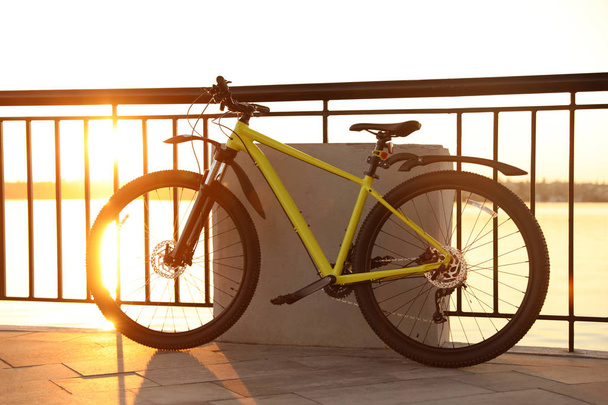Keltainen polkupyörä pysäköity lähelle kaidetta kaupungin rannalla auringonlaskun aikaan
 - Valokuva, kuva