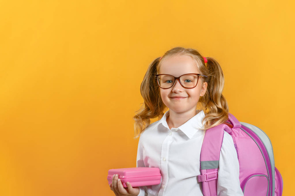 Portrait of a little girl students in glasses with a backpack and lunchbox on a yellow background. Back to school. The concept of education. Copy space - Photo, image