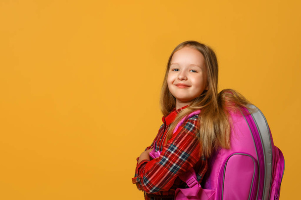 Portrait of a little girl schoolgirl on a yellow background. The child has a backpack behind his back. Back to school. The concept of education - Foto, imagen