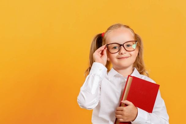 Portrait of a cute little girl on a yellow background. Child schoolgirl looking at the camera, holding a book and straightens glasses. The concept of education. Copy space - Foto, imagen