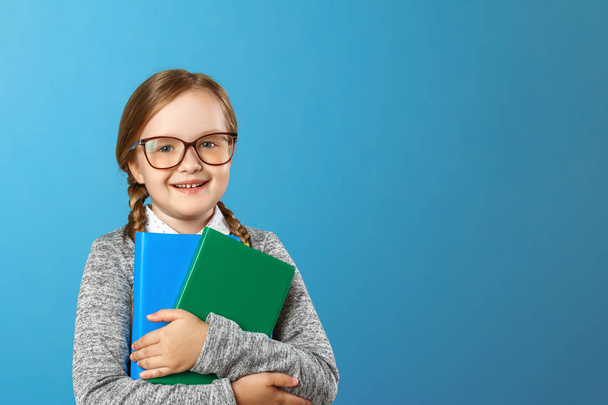 Portrait of a charming little girl schoolgirl in glasses on a blue background. The child holds books. The concept of education and school. Copy space - Foto, Imagen