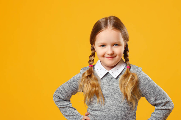 Portrait of a cute little girl on a yellow background. Child schoolgirl made hands to the side and looks into the camera. The concept of education - Φωτογραφία, εικόνα