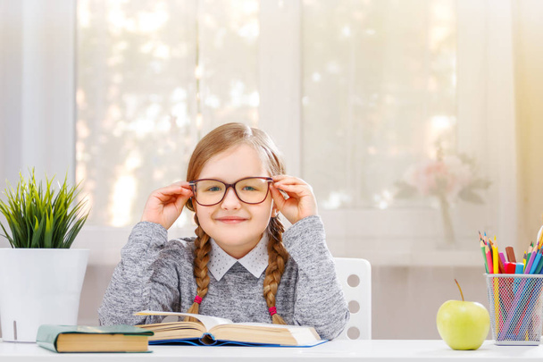Cute little student girl sitting at the table. The child straightens glasses and looks into the camera. The concept of education and school. Sunlight from the window. Blurred background. - Photo, Image
