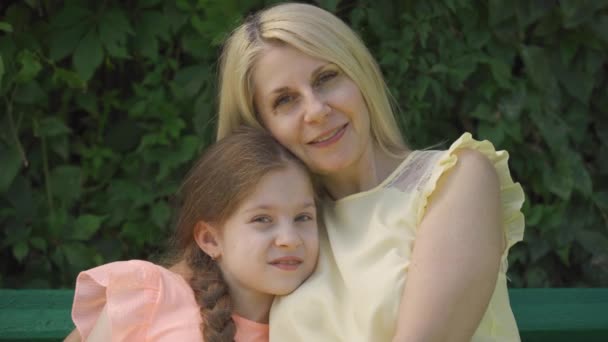 Portrait of mature blond mother in glasses and her little daughter looking at camera while sitting on the bench in the summer park and hugging. Happy family. Woman and girl together outdoors. - Metraje, vídeo