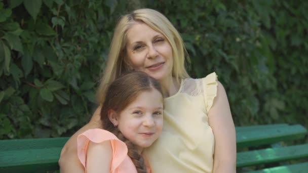 Portrait of pretty mature blond mother in glasses and her little daughter looking at camera while sitting on the bench in the summer park and hugging. Happy family. Woman and girl together outdoors. - Video