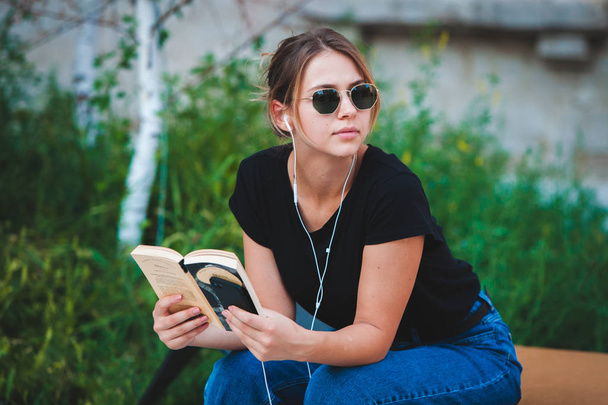 Ένα νεαρό κορίτσι που διαβάζει ένα βιβλίο στο δρόμο. γυναικεία χίπστερ που απολαμβάνει λογοτεχνία σε εξωτερικούς χώρους. Χαμογελαστός έφηβος κορίτσι ντυμένος σε απλό βιβλίο ανάγνωσης. - Φωτογραφία, εικόνα