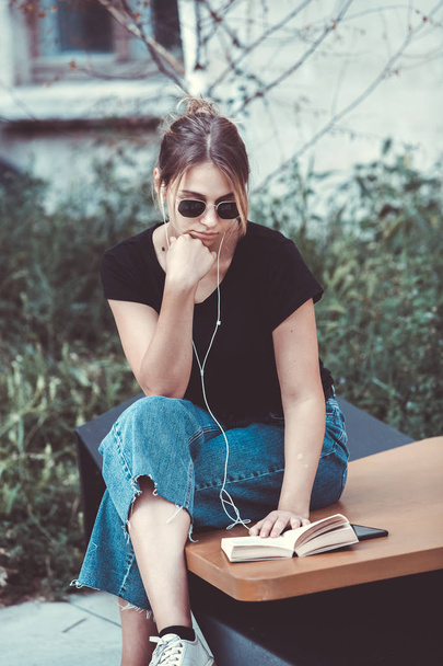 Young girl reading a book in the street.  female hipster enjoying literature outdoors. Smiling teen girl dressed in casual reading book. - Foto, immagini