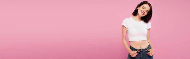panoramic shot of beautiful smiling girl with hands in pockets isolated on pink - Fotoğraf, Görsel