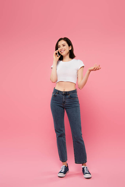 full length view of beautiful smiling girl talking on smartphone and gesturing isolated on pink - Foto, Imagem