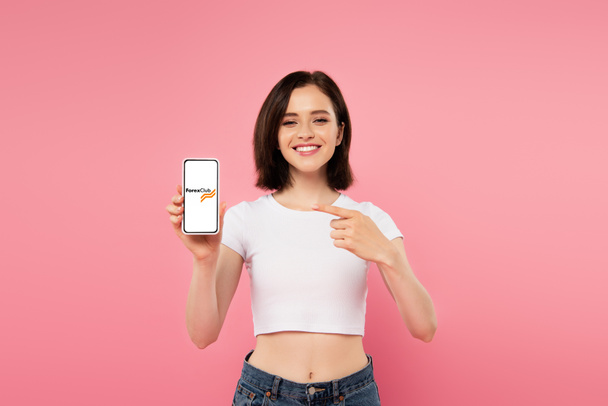 KYIV, UKRAINE - JULY 3, 2019: smiling girl pointing with finger at smartphone with Forex club logo isolated on pink - Foto, Imagen