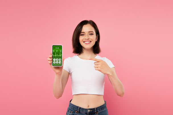 ragazza sorridente punta con il dito allo smartphone con app sanitaria isolata su rosa
 - Foto, immagini