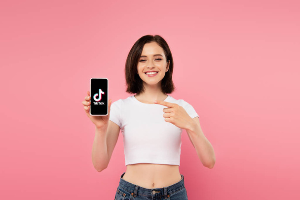 Київ, Україна-3 липня 2019: усміхнена дівчина вказуючи пальцем на смартфон з логотипом тіток, ізольованих на рожевому - Фото, зображення