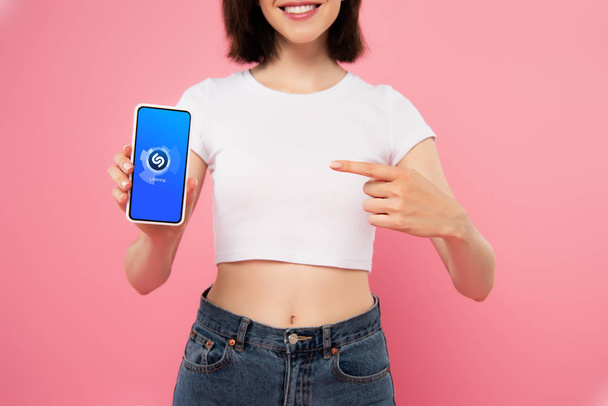 Київ, Україна-3 липня 2019: обрізаний вигляд дівчини вказуючи пальцем на смартфон з логотипом Shazam, ізольованих на рожевому - Фото, зображення
