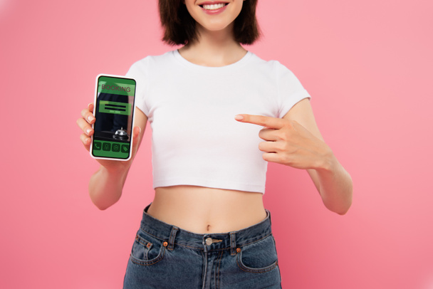 обрізаний вид дівчини, що вказує пальцем на смартфон із додатком бронювання ізольовано на рожевому
 - Фото, зображення