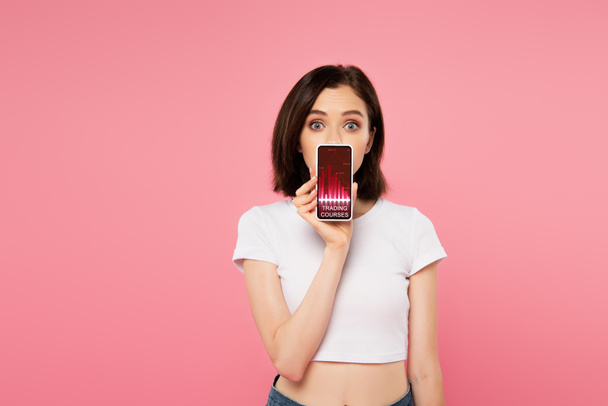 surprised girl holding smartphone with trading courses app isolated on pink - Photo, Image