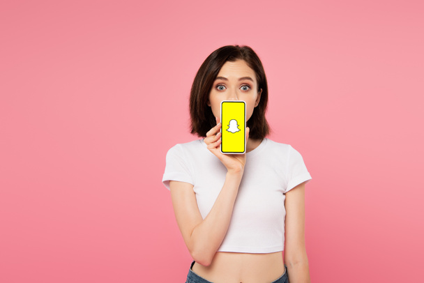 Kijev, Ukrajna-július 3, 2019: döbbent lány Holding smartphone snapchat logo izolált rózsaszín - Fotó, kép